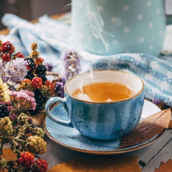 Димяща чаша чай в синя шарена чаша с чинийка, заобиколена от горски плодове и цветя върху дървена маса.