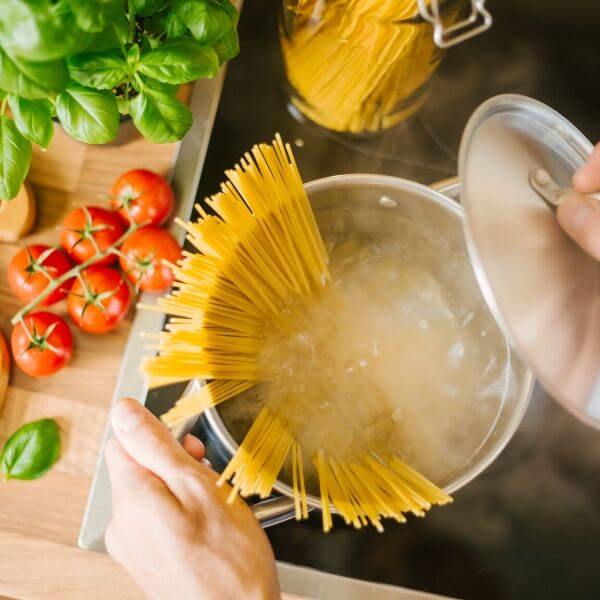 Човек готви спагети в тенджера с вряща вода, заобиколен от съставки като домати и босилек върху дървен плот.