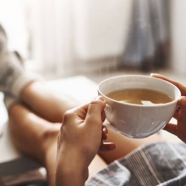 Човек в уютна обстановка, държащ голяма чаша чай, с частичен изглед към неговата спокойна поза при мека светлина.
