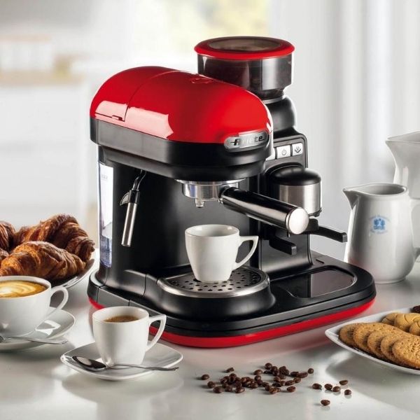 Червена и черна машина за еспресо с чаши кафе, кафе на зърна, кроасани и бисквити на кухненски плот.