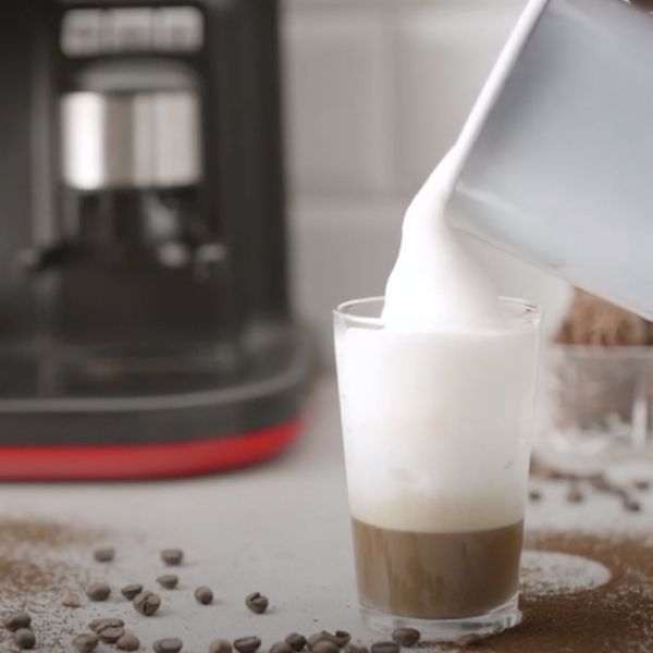 Мляко се налива в чаша с кафе на кухненски плот с разпръснати кафеени зърна и машина за еспресо на заден план.