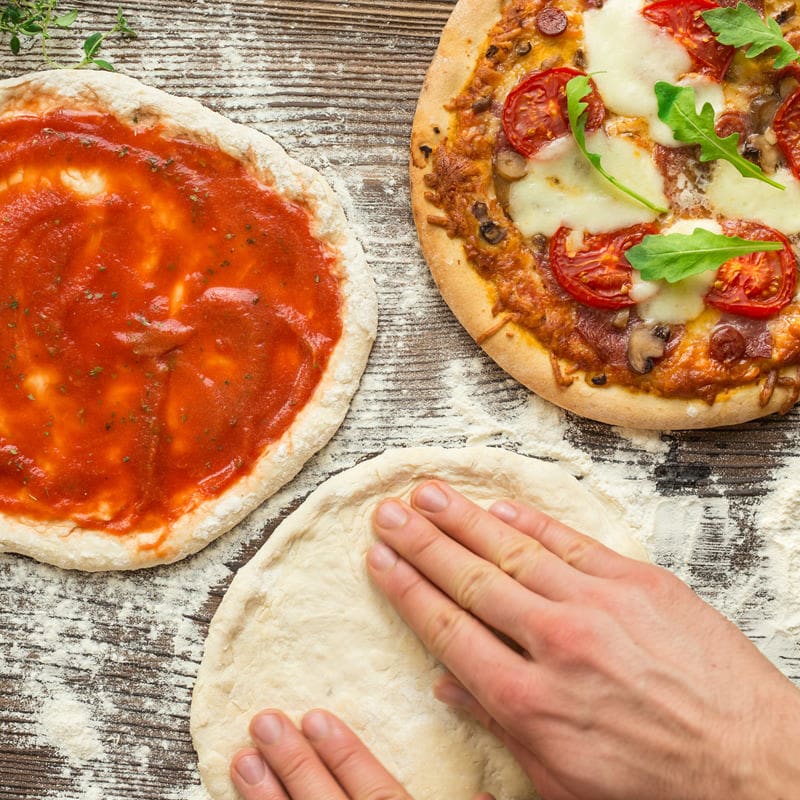Три етапа на приготвяне на пицата, показани върху дървена повърхност: тесто, разстилано на ръка, кора с доматен сос и изпечена пица, покрита със сирене, домати и билки с Хлебопекарна PANEXPRESS 1000.