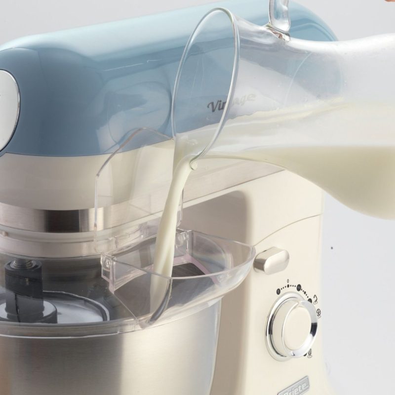Мляко се излива в прозрачна купа на кухненски робот от стъклена кана, до синьо-бял кухненски уред.