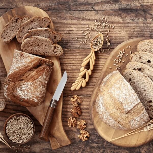 Различни видове нарязан хляб и цели хлябове върху дървени дъски с пшенични зърна, орехи и нож върху селска дървена маса.