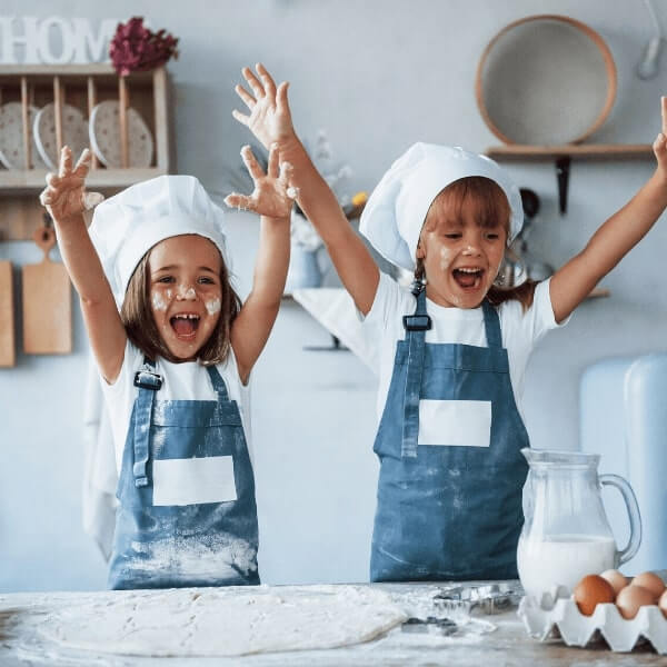 Две радостни деца с шапки и престилки на готвач вдигат ръце от вълнение, докато правят бъркотия с брашно в кухня.