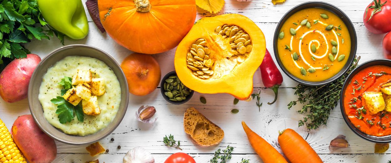 Ярко разнообразие от вдъхновени от есента ястия с разнообразни супи в купи, заобиколени от асортимент от пресни зеленчуци и билки върху дървена повърхност, перфектно смесени с помощта на Пасатор PIMMY 500 3 в 1