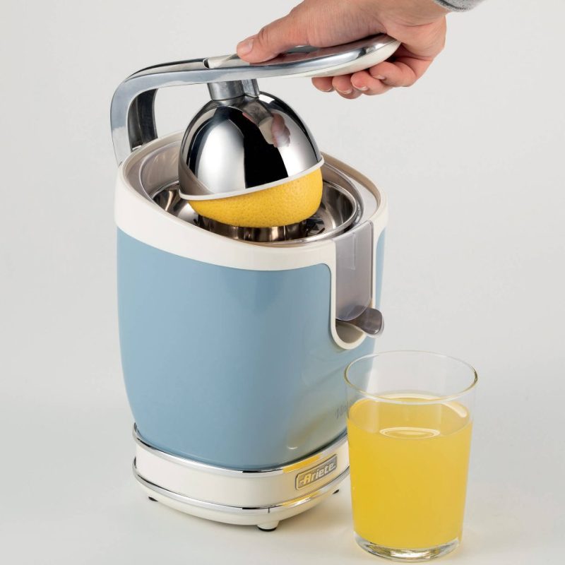Ръка на човек, използващ синя и сребриста електрическа сокоизстисквачка за цитрусови плодове, за да изцеди сок от нарязан наполовина лимон, с чаша пресен сок до него.