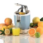 Електрическа сокоизстисквачка за цитрусови плодове, заобиколена от пресни портокали, лимони и чаша прясно изцеден портокалов сок на бял фон.