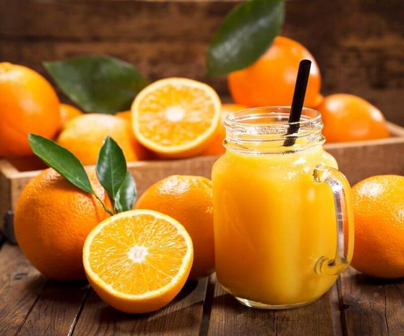 Пресен портокалов сок в буркан със сламка, заобиколен от цели и разполовени портокали върху дървена маса.