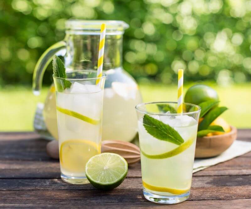 Две чаши лимонада със сламки и листа от мента, заедно с кана и пресен лайм, на дървена маса на открито.
