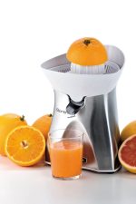 Електрическа Цитрус преса SPREMI METAL заобиколена от цели и разполовени портокали и нарязан грейпфрут, с чаша прясно изцеден портокалов сок на преден план.
