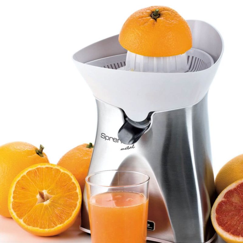 Електрическа Цитрус преса SPREMI METAL заобиколена от цели и разполовени портокали и нарязан грейпфрут, с чаша прясно изцеден портокалов сок на преден план.