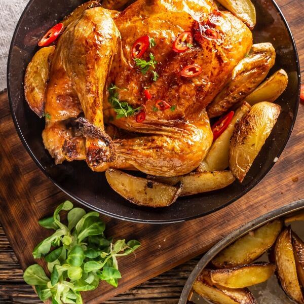 Печено пиле, гарнирано с червени люти чушки и билки, поднесено с печени картофи на селска дървена маса.