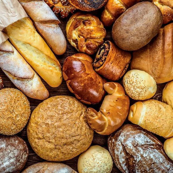 Асорти от прясно изпечени хлябове, включително багети, кифлички и хлябове, подредени плътно един до друг.