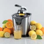 Цитрус преса PRO JUICE от неръждаема стомана, разпределяща пресен сок в чаша, заобиколена от цели портокали и лимони.