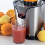 Цитрус преса PRO JUICE от неръждаема стомана с портокали и лимони около него, с чаша прясно изцеден сок на преден план.
