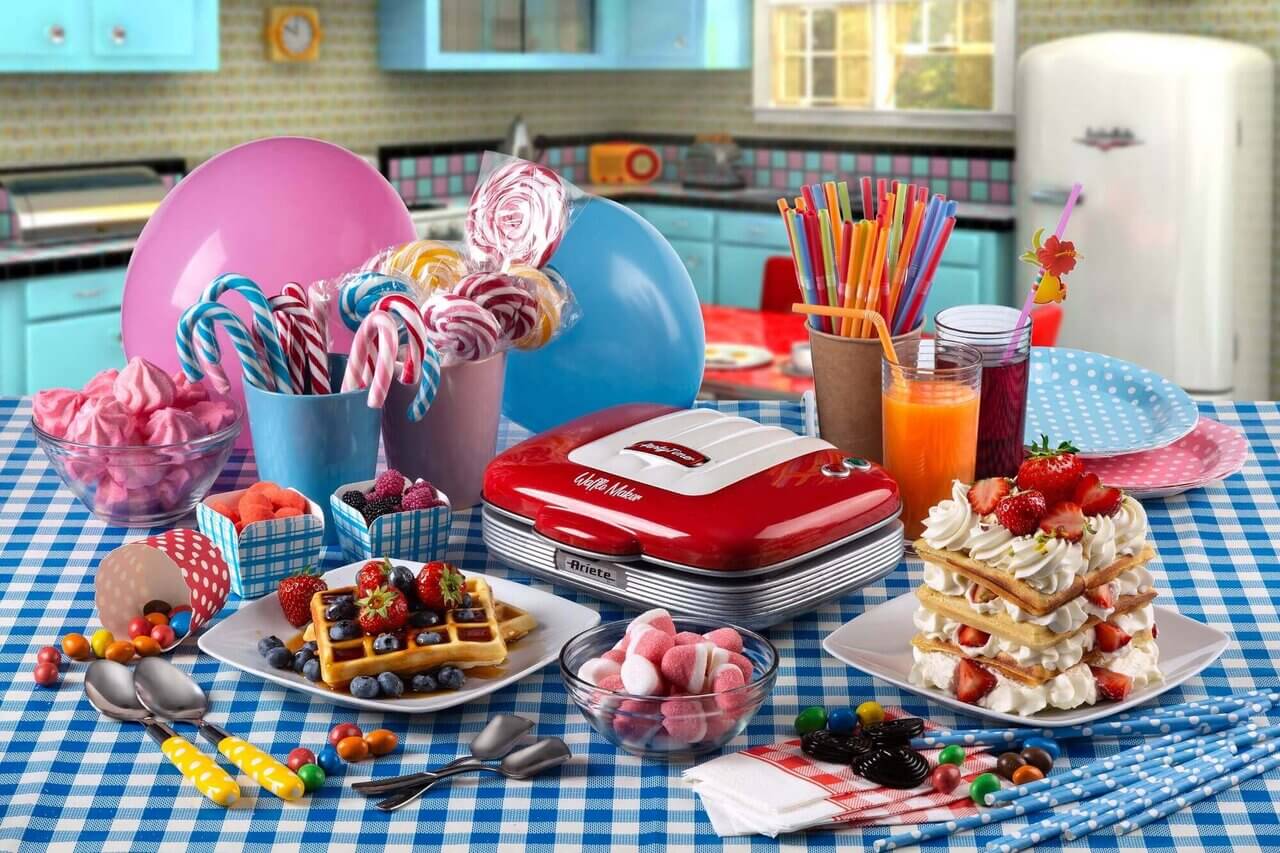 Цветна сцена със закуска с вафли, плодове, бонбони и напитки върху кухненска маса, украсена с балони и артикули за парти.