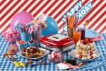 Цветни закуски и напитки за рожден ден с машина за гофрети, балони и надпис „Честит рожден ден“ върху карирана покривка.