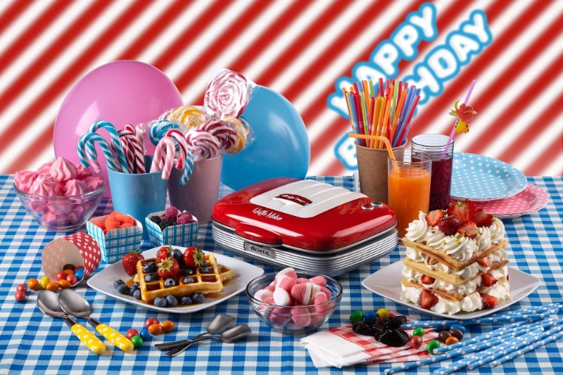 Цветни закуски и напитки за рожден ден с машина за гофрети, балони и надпис „Честит рожден ден“ върху карирана покривка.