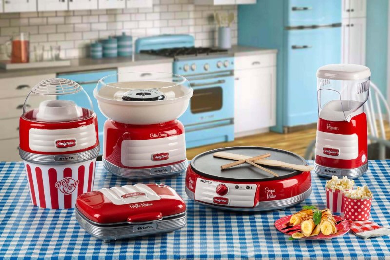 Различни червени и бели ретро кухненски уреди, включително машина за пуканки, мини грил и машина за креп, изложени върху синя карирана покривка.