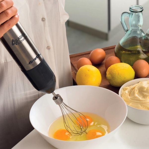 Човек използва Мултифункционален пасатор 7в1 - 4 ОСТРИТА, за да разбие яйца в бяла купа, със съставки като лимони, яйца и бутилка олио, поставени наблизо на кухненския плот.