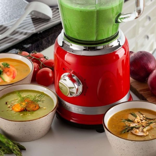 Червен блендер MODERNA със зелена смес стои на плота, заобиколен от пресни съставки и купи с кремообразни супи.