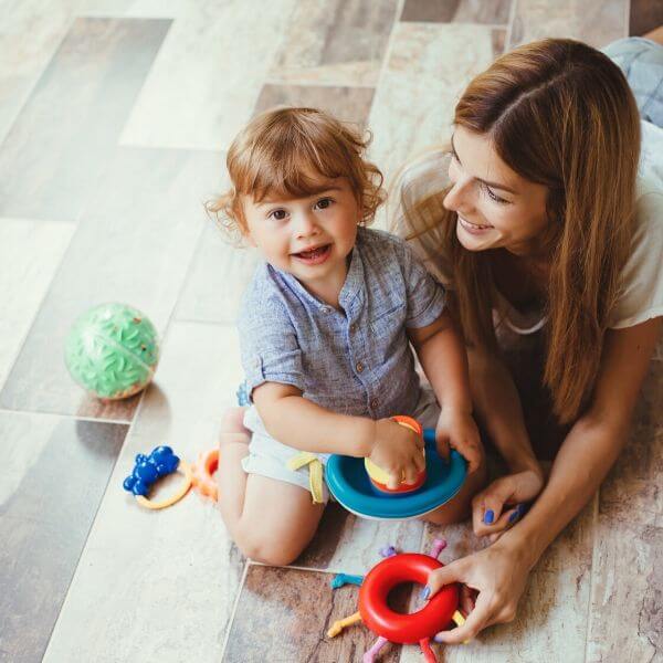 Майка и дете играят с играчки на пода. Майката демонстрира своите Вертикална прахосмукачка HANDY FORCE умения, безпроблемно маневрирайки около.