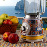 Блендерът VINTAGE със стъклена кана – Positano стои на маса с плодове, откъдето се разкрива зашеметяваща гледка към морето.
