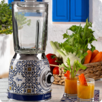 Блендерът VINTAGE със стъклена кана – Capri стои на масата до две чаши сок и кошница със зеленчуци.