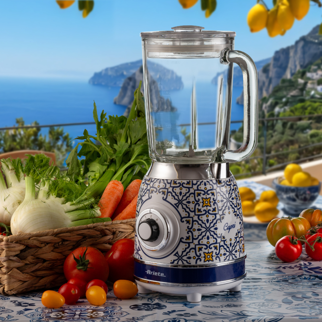 Блендерът VINTAGE със стъклена кана – Capri стои на маса със зеленчуци на балкон с изглед към морето и хълмовете.