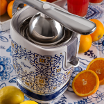 Цитрусова преса VINTAGE JUICER – Capri със синьо-жълт мотив, заобиколена от портокали и лимони върху шарена кърпа.
