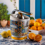 Цитрусопресата VINTAGE JUICER – Positano е поставена върху маса с мотиви, върху която има лимони, портокали и кутия с цитрусови плодове.