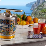 Цитрусова преса VINTAGE JUICER – „Позитано“ с пресни плодове и сок, поставена на маса с изглед към крайбрежната скала.