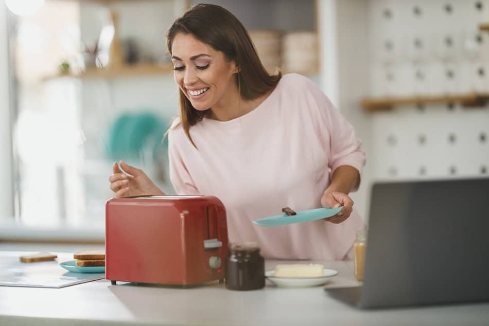 Жена се усмихва, докато прави тост с червения си тостер Ariete, държи чиния и нож за масло близо до лаптоп.