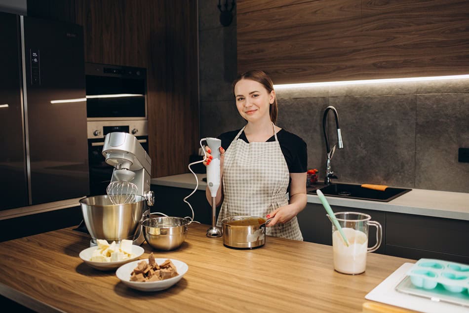 Жена с престилка използва ръчен миксер Ariete в модерна кухня, а на плота са поставени различни съставки за печене.