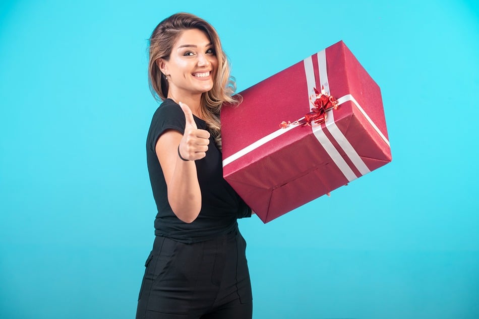 Жена, която държи червена кутия за подаръци Ariete с бяла панделка и вдига палец на син фон.