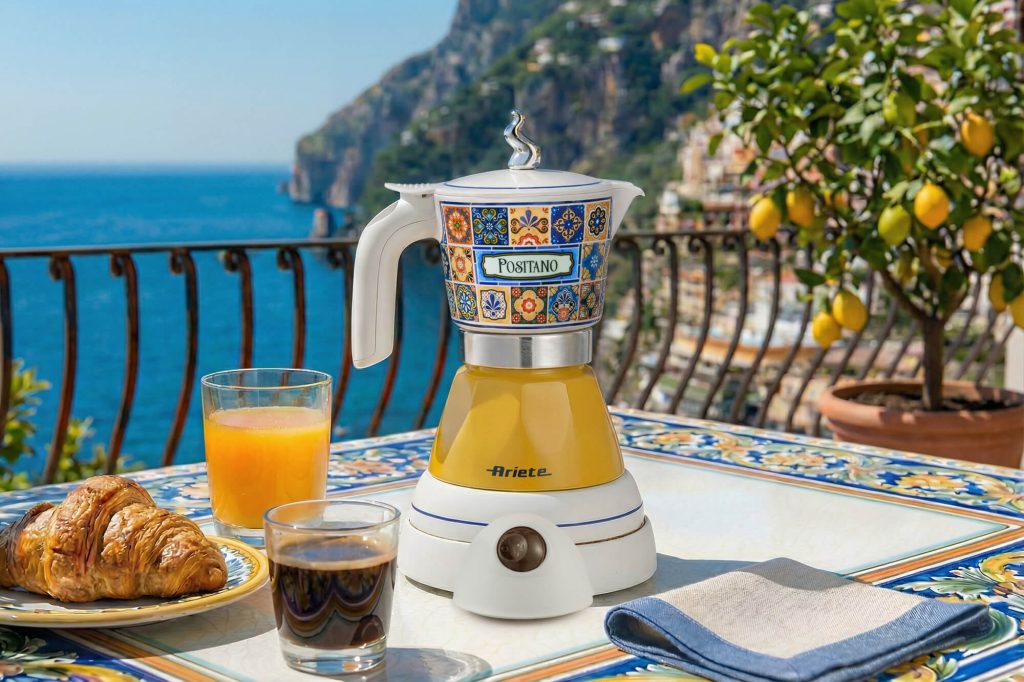 Електрическа кафемашина „Moka Vintage Positano“, сок и кроасан на плочките край морето и скалите.