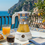 Електрическа кафемашина „Moka Vintage Positano“ върху плочки, до чаша кафе, сок и кроасан край морето.