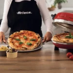 Някой, облечен в престилка на Ariete, държи пица от Фурна за пица "Pizzeria Gourmet", с маслини и артишок наблизо.