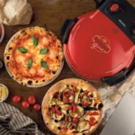 Две пици на маса до Фурна за пица "Pizzeria Gourmet", пресни домати, тесто и кухненски прибори.