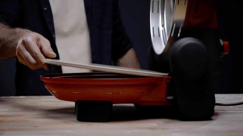 Човек използва дървена кора, за да вкара пица в червената Фурна за пица "Пицария Гурме".