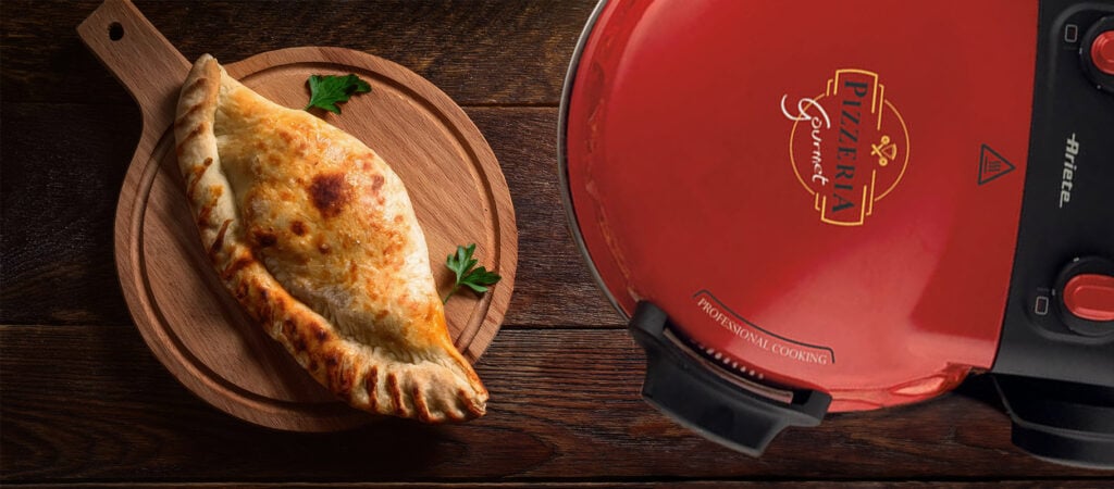 Изпечена калцоне върху дървена чиния до червена Фурна за пица "Pizzeria Gourmet" върху дървена маса.