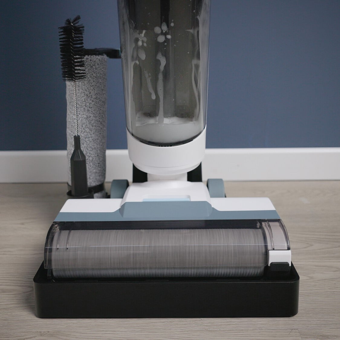 Близък план на Вертикална прахосмукачка Aqua Force Wash & Vacuum с ролкова четка върху дървен под, синя стена отзад.