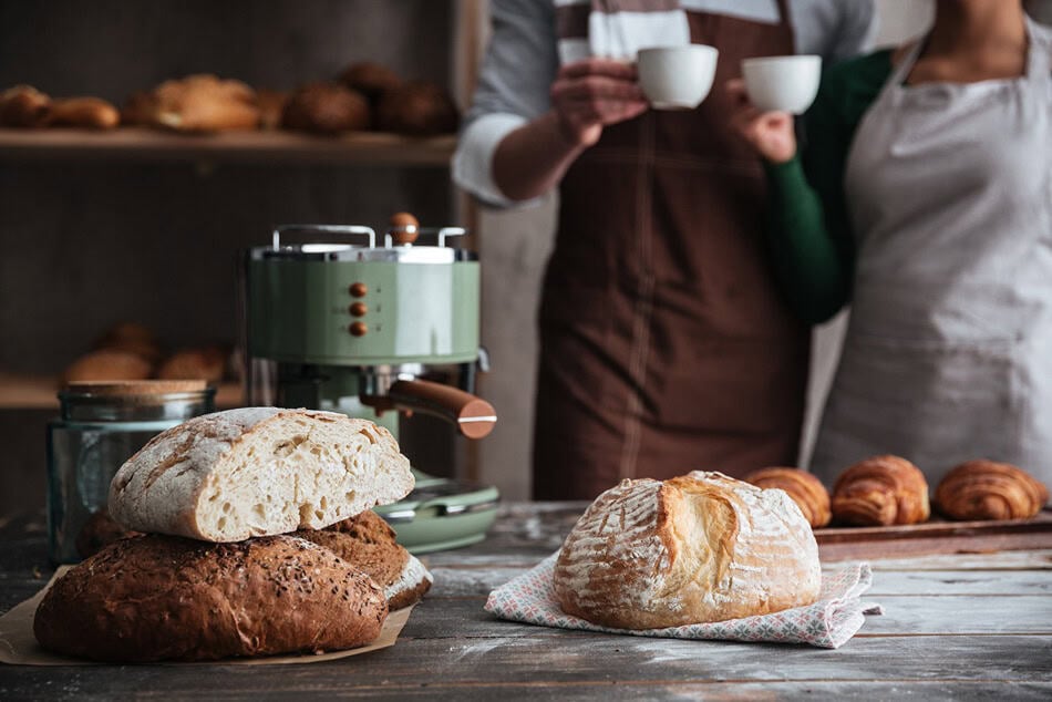 Двама души в престилки стоят с чаши за кафе зад хлебопекарна с пресен хляб и зелена машина за еспресо.