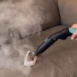 Една ръка почиства сив тапициран диван с парочистачка X-Vapor Comfort, 5 бара и кърпа.