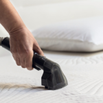 Човек почиства бял матрак с уреда за премахване на петна Spot Cleaner Heat Pro, а зад него има бяла възглавница.