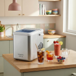 Сребрист ледогенератор „Ice Machine“ с капацитет 12 кг/24 ч. е поставен на кухненския плот, заобиколен от ледени напитки, купа с горски плодове и чаша с лед, а на заден план се виждат кухненски рафтове и слънчева светлина.