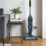Прахосмукачката за мокро и сухо почистване AquaForce Easy Wash & Vacuum се зарежда от контакт до малка странична масичка с книги и саксийно растение, разположена до сив диван върху дървен под. Наблизо стои автоматична машина за сладолед, готова да приготви вкусни лакомства, което придава съвременно удобство на уютното жилищно пространство.
