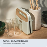 Компактен, модерен кухненски уред стои изправен върху дървен плот, заобиколен от чаши, зелени купички и бежова ваза с дървени прибори. Текстът на български език описва неговия тънък и компактен дизайн, подходящ за всяка кухня.