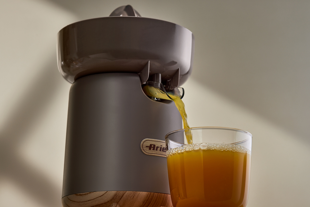 Цитрусовата преса „Breakfast Wood“, с металично сиво покритие и дървена основа, плавно излива пресен портокалов сок в прозрачната чаша под нея.